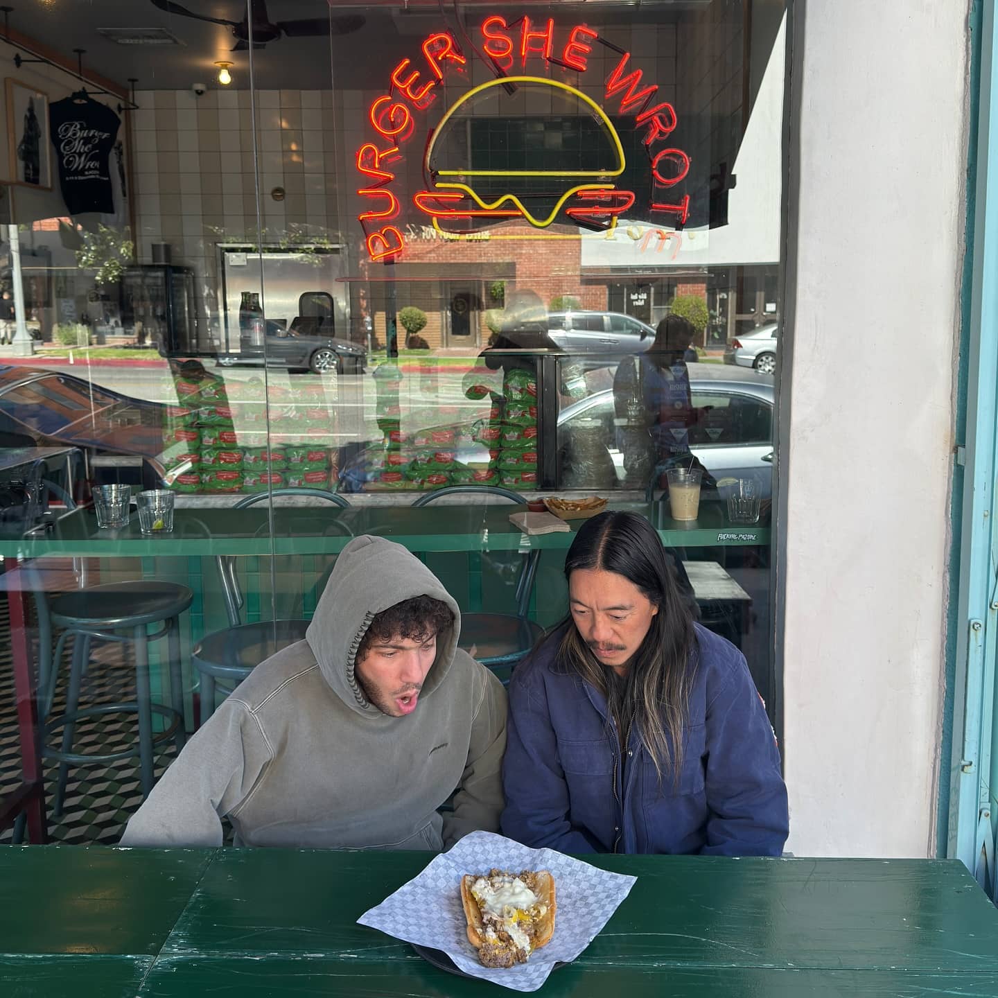 Two people sharing a burger at the window counter under the Burger She Wrote neon sign